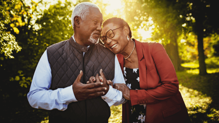 A happy senior couple, with the woman resting her head on the man's shoulder and holding his hand, stands in a sunlit park, representing a fulfilling lifestyle and spending in retirement, which is a goal of good retirement planning.